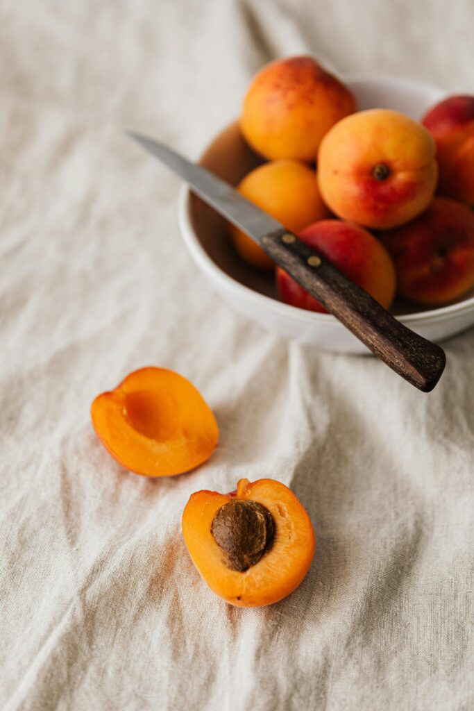 From above of ceramic bowl with ripe colorful fruits and knife placed on white rumpled tablecloth near cut peach in half